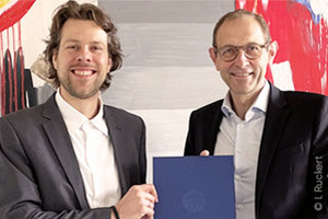 Jun. Prof Raoul van Maarseveen and WiSo Dean Prof. Ulrich Thonemann Ph.D., smiling in front of an abstract painting, together holding a blue folder with the logo of the University of Cologne in the camera. 