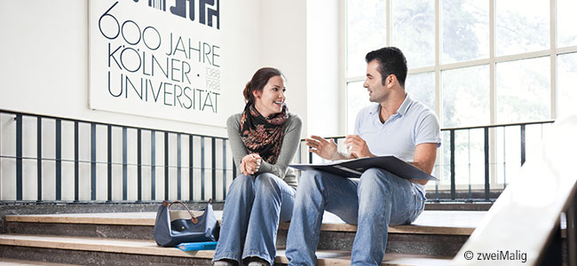Eine Studentin und ein Student sitzen auf einer Treppe und unterhalten sich, dahinter der Schriftzug auf einem Bild: 600 Jahre Kölner Universität